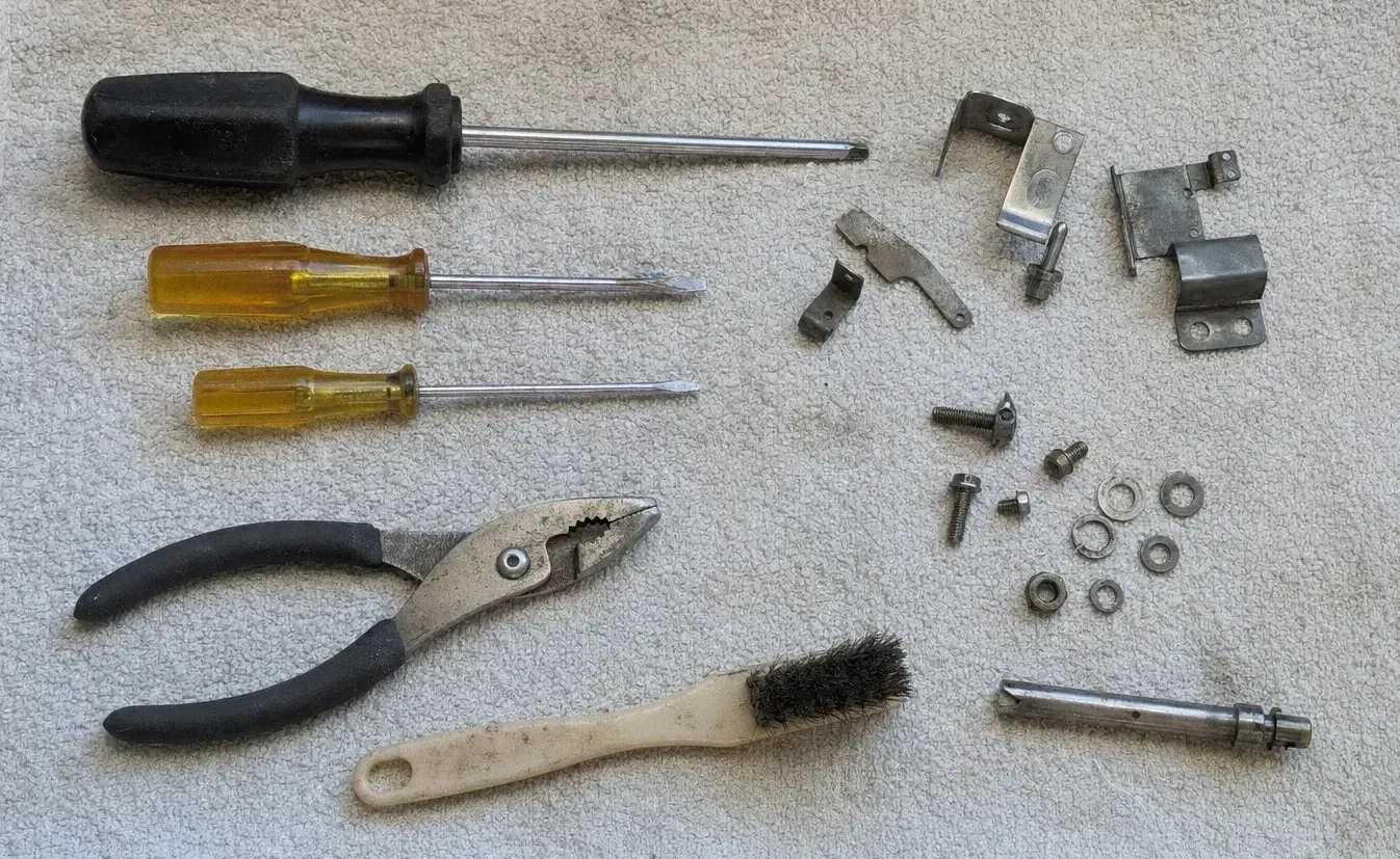 Three screwdrivers, a long shaft driver, slip-joint pliers and a wire brush spread on a cloth beside hinge brackets, washers and screws